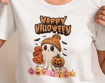 May include: White t-shirt with a Halloween-themed graphic. The design features a friendly ghost wearing a knit cap, surrounded by carved pumpkins and bats. The words "Happy Halloween" are printed above the ghost in orange letters.