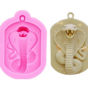 以下が含まれることがあります： コブラのペンダントを作るためのピンクのシリコンモールド。モールドは、フードを立てて口を開けたコブラの形をしています。モールドは、モールドから作られた完成したペンダントの隣に示されています。