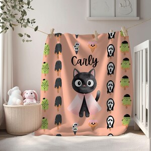 Könnte beinhalten: Eine pfirsichfarbene Decke mit einer schwarzen Katze mit rosa Umhang, dem Namen "Carly" und Halloween-Eiscreme- und Geistermotiven. Die Decke hängt an einer Wäscheleine mit Wäscheklammern aus Holz.