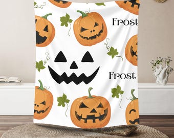 Op de afbeelding: Witte Halloween-deken met oranje pompoengezichten, groene bladeren en het woord "Frost" in zwart. De deken is gedrapeerd over een rond, geweven tapijt.