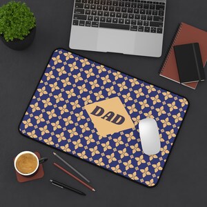 Pode incluir: Um mousepad retangular com um padrão floral azul e a palavra "DAD" em um retângulo dourado. Um mouse branco está no mousepad, com um laptop, um caderno e uma xícara de café no fundo.