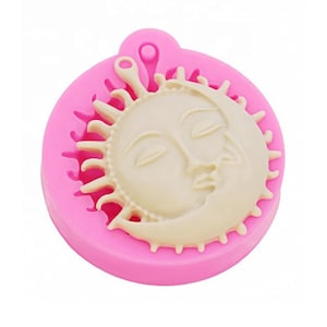 May include: Pink silicone mold for making a sun and moon pendant. The mold is round and has a raised design of a sun with a face and a crescent moon.