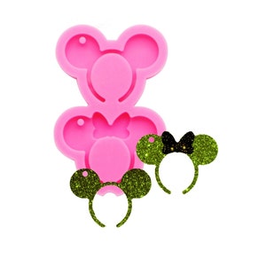 May include: Pink silicone mould for making resin mouse ear headbands. The mould has two cavities, one for a larger headband and one for a smaller headband. The mould is shown with two finished headbands, one with green glitter and a black bow, and one with green glitter and a black bow.