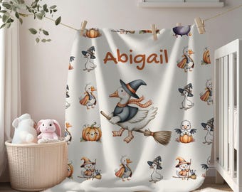 Könnte beinhalten: Eine weiße Babydecke mit Halloween-Motiv. Die Decke zeigt Cartoon-Gänse in Hexenkostümen, Kürbisse und den Namen "Abigail" in Orange. Die Decke hängt an einer Wäscheleine und ist eine charmante Dekoration für das Kinderzimmer.