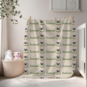 May include: A beige blanket with a repeating pattern of pug dog illustrations, yellow flowers, and the name "Samuel" in green script. The blanket is hanging on a clothesline in a nursery setting.
