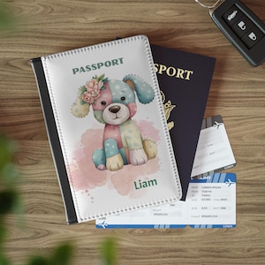 Puede incluir: Una funda blanca para pasaporte con una ilustración de perro de retazos de colores, el nombre "Liam" en verde y la palabra "PASSPORT" en verde. También se ven un pasaporte azul oscuro, billetes de avión y una llave de coche.