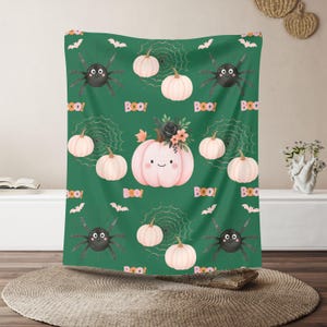 Könnte beinhalten: Eine grüne Halloween-Decke mit einem sich wiederholenden Muster aus lächelnden rosa Kürbissen, schwarzen Spinnen, Spinnweben und Fledermäusen. Das Wort "Boo!" ist wiederholt. Die Decke ist aus weichem Material.