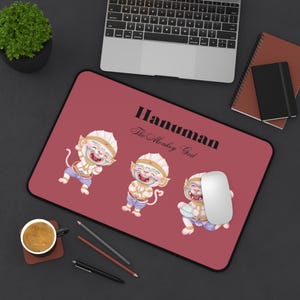Puede incluir: Una alfombrilla de ratón roja con tres ilustraciones de dibujos animados de Hanuman, el Dios Mono, y el texto "Hanuman The Monkey God" en negro. Un ratón de ordenador blanco descansa sobre la alfombrilla, con un portátil y otros útiles de oficina en el fondo.