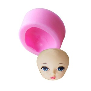 Peut inclure: Moule en silicone rose pour faire une tête de poupée. Le moule est ouvert et une tête de poupée finie est présentée à côté. La tête de poupée est faite d'un matériau brun clair et a des yeux bleus, une bouche rose et des sourcils peints.