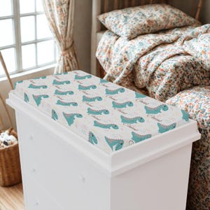 May include: A white changing pad cover with a repeating pattern of teal dinosaurs and the word "Caius" in a script font. The dinosaurs are surrounded by small, colorful confetti-like shapes.
