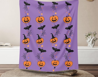 Op de afbeelding: Een paars Halloween-tapijt met een herhalend patroon van oranje pompoenen, zwarte vleermuizen, spinnenwebben en sterren. De pompoenen hebben lachende gezichten en zwarte stelen. De vleermuizen vliegen en de sterren zijn geel.
