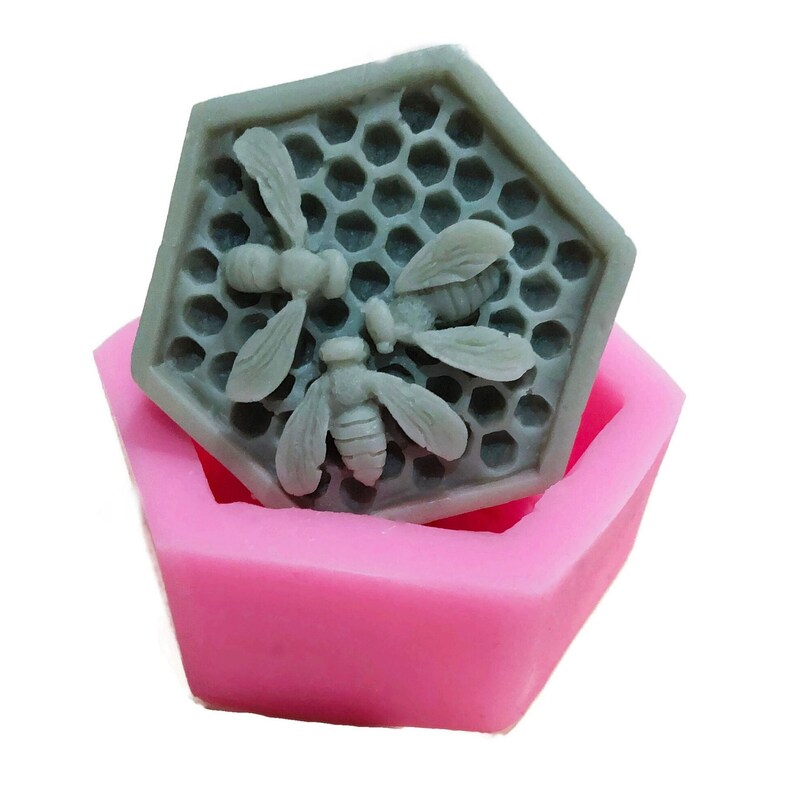 Może przedstawiać: R&oacute;żowa silikonowa forma w kształcie sześciokąta z szarą sześciokątną mydłem w środku. Mydło ma wz&oacute;r plastra miodu i trzy pszczoły.