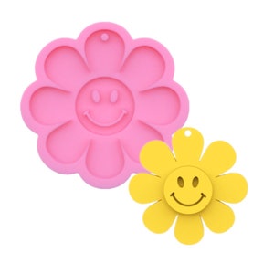 Puede incluir: Molde de silicona rosa en forma de flor con una cara sonriente en el centro. Una flor de resina amarilla con una cara sonriente se muestra junto al molde.