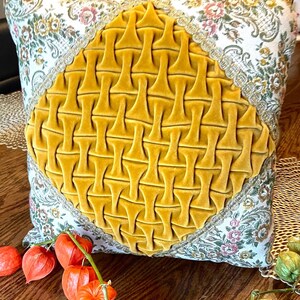 May include: Square decorative pillow with a mustard yellow velvet center design. The pillow has a floral pattern on a cream background with gold trim. The pillow is on a wooden surface with orange flowers.