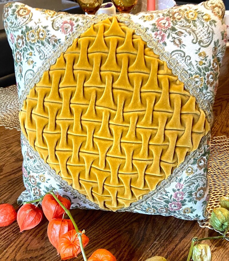 May include: Square decorative pillow with a mustard yellow velvet center design. The pillow has a floral pattern on a cream background with gold trim. The pillow is on a wooden surface with orange flowers.