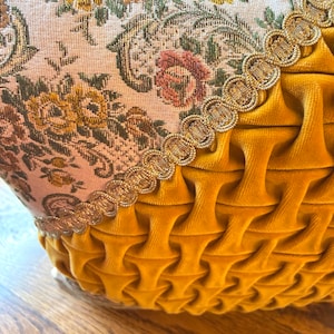 May include: Close-up of a decorative pillow with a floral tapestry top and a mustard yellow velvet bottom. The tapestry features a cream background with pink, yellow, and green floral patterns. A gold trim separates the two fabrics.
