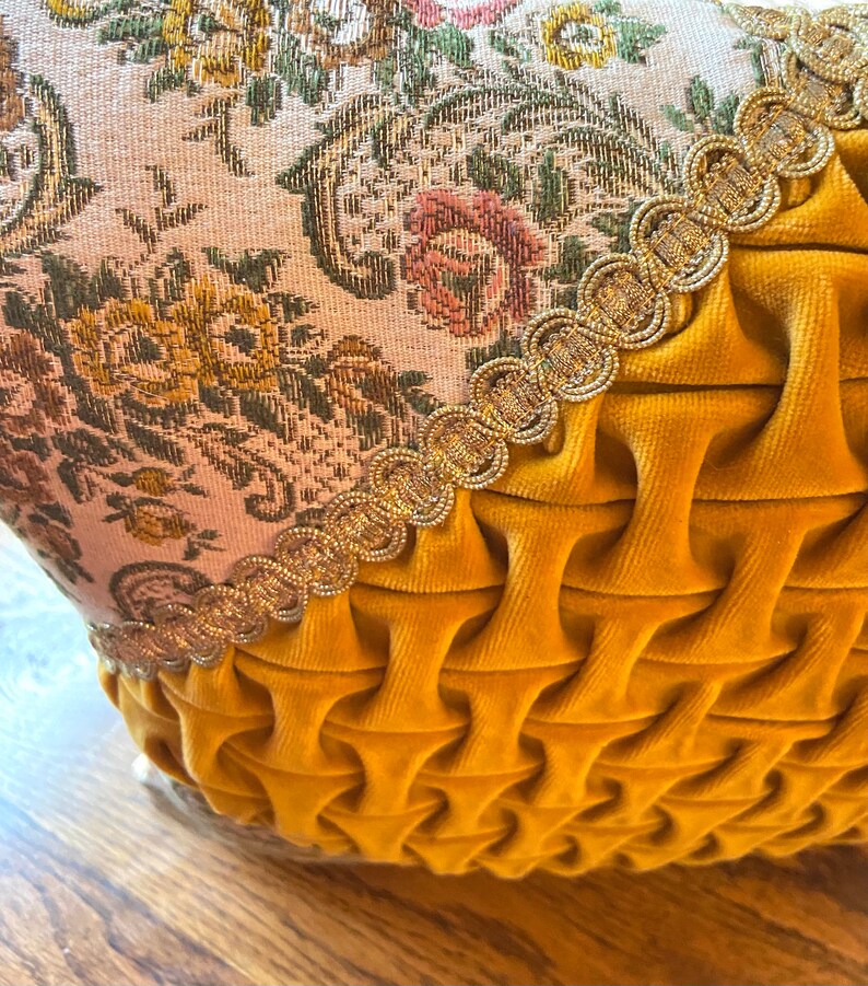 May include: Close-up of a decorative pillow with a floral tapestry top and a mustard yellow velvet bottom. The tapestry features a cream background with pink, yellow, and green floral patterns. A gold trim separates the two fabrics.