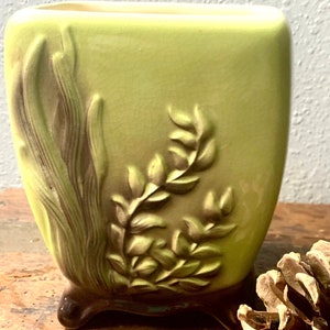May include: A vintage green ceramic planter with a raised floral design. The planter has a square shape with rounded edges and three small feet.