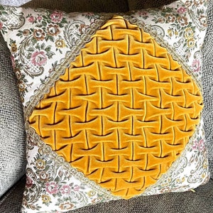 May include: Square decorative pillow with a yellow velvet center featuring a woven design. The pillow has floral patterned fabric on the sides with a gold trim. The pillow is on a gray surface.