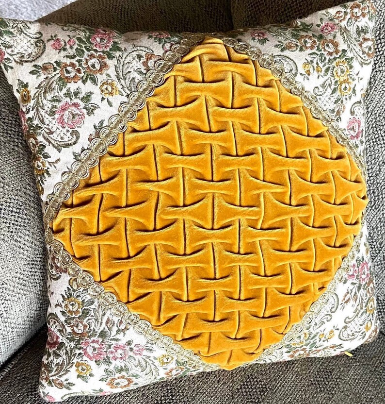 May include: Square decorative pillow with a yellow velvet center featuring a woven design. The pillow has floral patterned fabric on the sides with a gold trim. The pillow is on a gray surface.