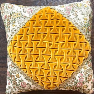 May include: Square decorative pillow with a floral patterned border and a central diamond-shaped design. The diamond is made of a textured, golden-yellow velvet fabric. The border features a cream-colored fabric with pink and yellow floral patterns.