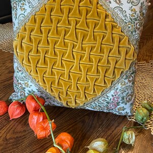 May include: Square decorative pillow with a mustard yellow velvet center and floral patterned fabric border. The pillow has a diamond-shaped, textured design in the center. Orange and green dried flowers are arranged on a wooden surface near the pillow.