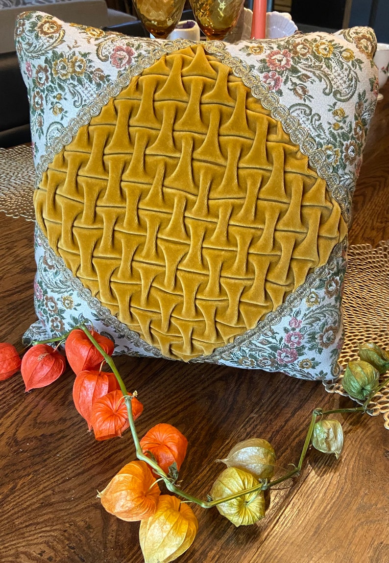 May include: Square decorative pillow with a mustard yellow velvet center and floral patterned fabric border. The pillow has a diamond-shaped, textured design in the center. Orange and green dried flowers are arranged on a wooden surface near the pillow.