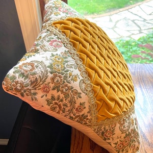 May include: Decorative pillow with a mustard yellow, woven, textured center and floral patterned sides. The sides feature a cream background with a floral design in shades of green, pink, and brown. Gold trim borders the yellow center.