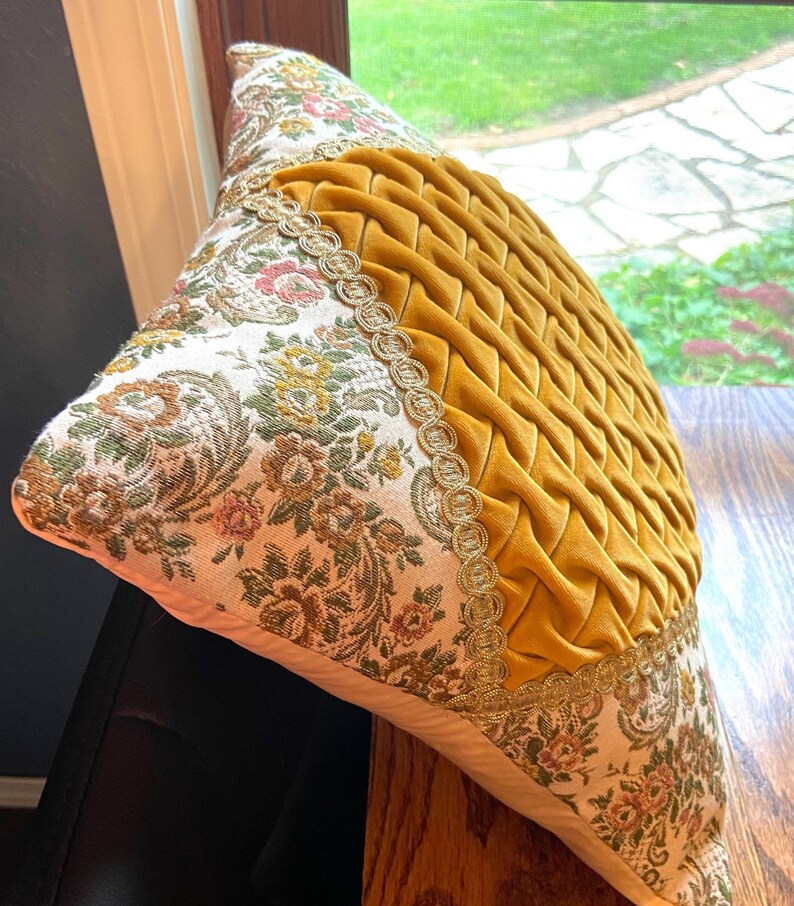 May include: Decorative pillow with a mustard yellow, woven, textured center and floral patterned sides. The sides feature a cream background with a floral design in shades of green, pink, and brown. Gold trim borders the yellow center.