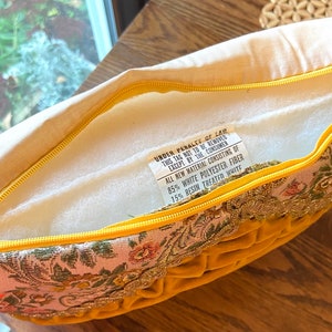 May include: A decorative pillow with a yellow velvet front and a floral patterned border. The pillow has a yellow zipper and a tag with text that reads "UNDER PENALTY OF LAW THIS TAG NOT TO BE REMOVED EXCEPT BY THE CONSUMER".