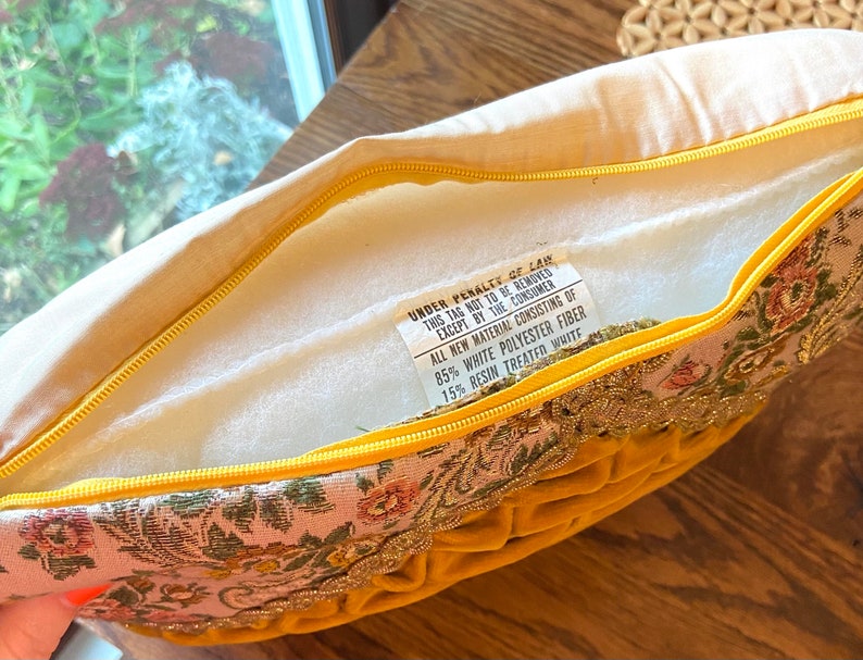 May include: A decorative pillow with a yellow velvet front and a floral patterned border. The pillow has a yellow zipper and a tag with text that reads "UNDER PENALTY OF LAW THIS TAG NOT TO BE REMOVED EXCEPT BY THE CONSUMER".
