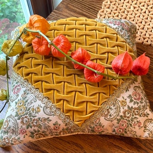 May include: Mustard yellow decorative pillow with a woven texture in the center. The pillow has floral patterned fabric on the edges, accented with gold trim. A stem of orange and green flowers rests on the pillow.