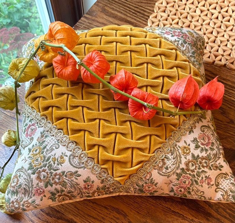 May include: Mustard yellow decorative pillow with a woven texture in the center. The pillow has floral patterned fabric on the edges, accented with gold trim. A stem of orange and green flowers rests on the pillow.