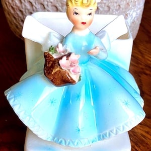May include: A vintage porcelain figurine of a seated woman in a light blue dress, holding a bouquet of pink flowers. The figurine has blonde hair and is set against a white backdrop with a bow. The dress has a scalloped hemline.