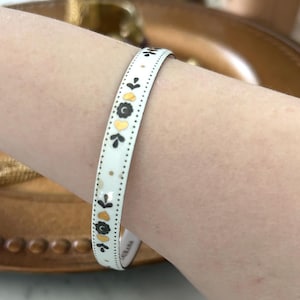 May include: White enamel bangle bracelet with a repeating pattern of black, gold, and yellow floral and leaf designs. The bracelet has a dotted border and is worn on a wrist. The bracelet is a decorative accessory.
