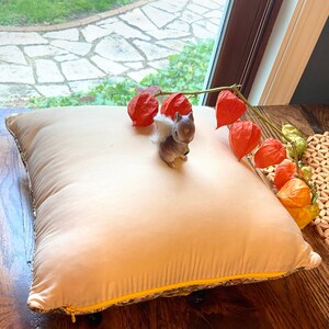 May include: A beige square pillow with a yellow zipper and floral trim. A small gray squirrel figurine with a white tail sits on the pillow. Orange Chinese lantern flowers are arranged nearby, creating a cozy, autumnal scene.