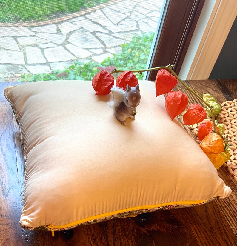 May include: A beige square pillow with a yellow zipper and floral trim. A small gray squirrel figurine with a white tail sits on the pillow. Orange Chinese lantern flowers are arranged nearby, creating a cozy, autumnal scene.