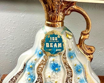 May include: A white ceramic decanter with a gold-colored top and handle. The decanter is decorated with blue and gold floral designs. The label on the front reads "155 Months Old Beam 4/5 Quart 86 Proof".