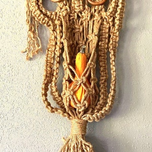 May include: A macrame wall hanging with a small orange gourd in the center. The hanging is made of natural jute rope and has a fringe at the bottom.