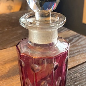 May include: A clear glass bottle with a faceted stopper. The bottle is a deep pink color and has a faceted design.