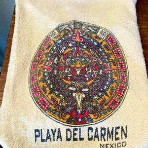 May include: A white canvas tote bag with a colorful Mayan calendar design and the text "PLAYA DEL CARMEN MEXICO".