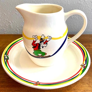 May include: White ceramic pitcher with a colorful cartoon of Goofy on it. The pitcher has a handle and is sitting on a matching saucer with a colorful stripe design.