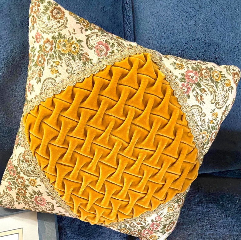 May include: Square decorative pillow with a floral patterned fabric border and a central section of pleated, mustard-yellow velvet. The pillow has a gold trim and is set against a blue background.