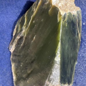 May include: A rough-cut, raw jade stone with a deep green hue. The stone has a natural, unpolished appearance with a textured surface and a lighter, beige-colored edge. The jade is displayed against a blue background.