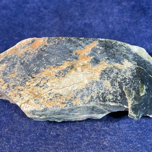 May include: A dark gray and green rock with a rough, uneven surface. The rock has a light brown and white pattern throughout.
