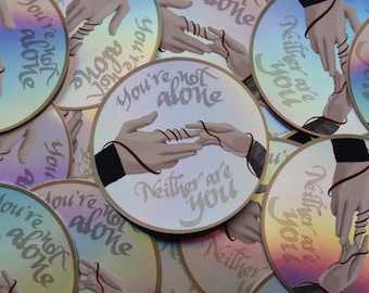 Könnte beinhalten: Holografische Aufkleber mit einem Design von zwei Händen, die durch einen roten Faden verbunden sind. Der Text "You're not alone" und "Neither are you" ist auf den Aufklebern geschrieben. Die Aufkleber haben ein reflektierendes, irisierendes Finish.