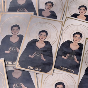 Könnte beinhalten: Eine Sammlung von Tarotkarten mit goldenem Rand und der Aufschrift "THE SUN". Jede Karte zeigt eine Illustration einer Frau in einem dunklen Kleid, die ein Sonnensymbol hält. Der Hintergrund ist hellgrau.
