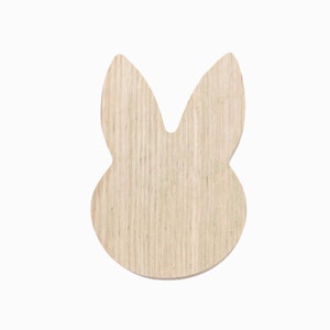 May include: A wooden bunny head cutout with a light brown, natural wood grain finish. The bunny shape features long ears and a rounded head, ideal for Easter crafts or home decor. The cutout is on a white background.