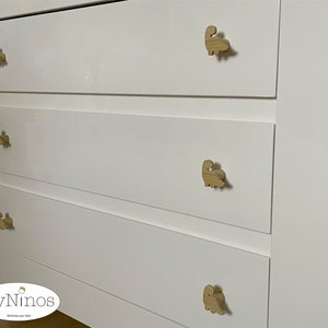 Op de afbeelding: Witte commode met vijf laden met houten knoppen in de vorm van een dinosaurus. De commode heeft een witte afwerking en de knoppen zijn van natuurlijke houtkleur.