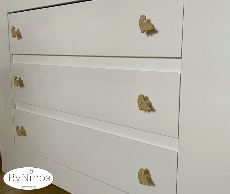 Op de afbeelding: Witte commode met vijf laden met houten knoppen in de vorm van een dinosaurus. De commode heeft een witte afwerking en de knoppen zijn van natuurlijke houtkleur.